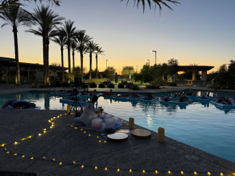 Sunset resort pool wellness scene with tall palm trees, people floating on blue mats during a twilight sound-bath session, musician with crystal bowls on the deck and warm string lights.