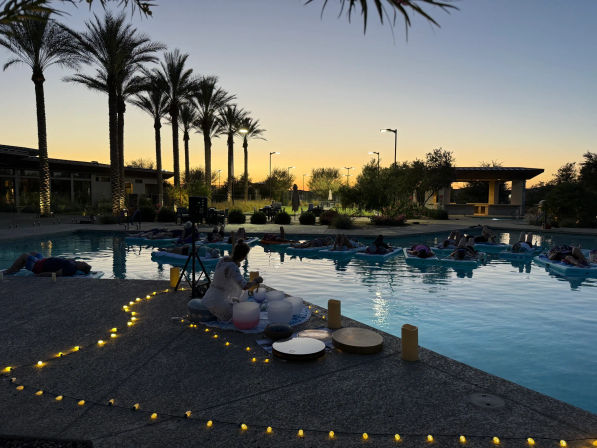 Sunset resort pool wellness scene with tall palm trees, people floating on blue mats during a twilight sound-bath session, musician with crystal bowls on the deck and warm string lights.