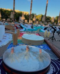 Outdoor poolside percussion setup with frame drums, shakers, microphone and candles on colorful mats, palm trees and lounge chairs in the background and palm reflections on a drum surface.
