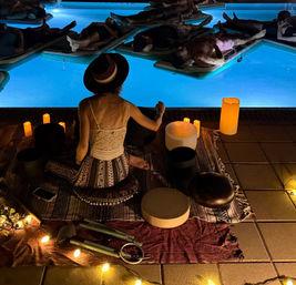 Nighttime outdoor poolside sound bath — person wearing a hat seated on a patterned rug playing singing bowls amid candles and string lights while participants float on mats in an illuminated blue pool for a relaxing meditation session.