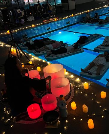 Evening indoor pool sound bath — people lying on floating mats in a glowing blue lap pool, circled by twinkle lights and LED candles while a musician plays illuminated crystal singing bowls on the deck.