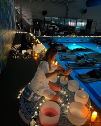 Evening indoor pool sound bath and meditation: a facilitator kneeling on a patterned mat with crystal singing bowls, candles and string lights while participants lie on floating mats in a softly lit blue pool during a wellness event.