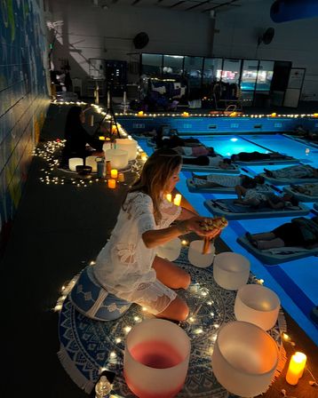Evening indoor pool sound bath and meditation: a facilitator kneeling on a patterned mat with crystal singing bowls, candles and string lights while participants lie on floating mats in a softly lit blue pool during a wellness event.