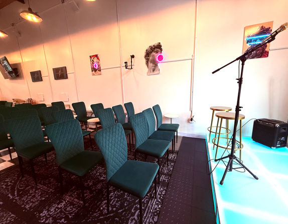 Intimate indoor event space with teal quilted chairs in rows facing a small illuminated stage with microphone stand, two gold stools, amp and neon-accent modern wall art
