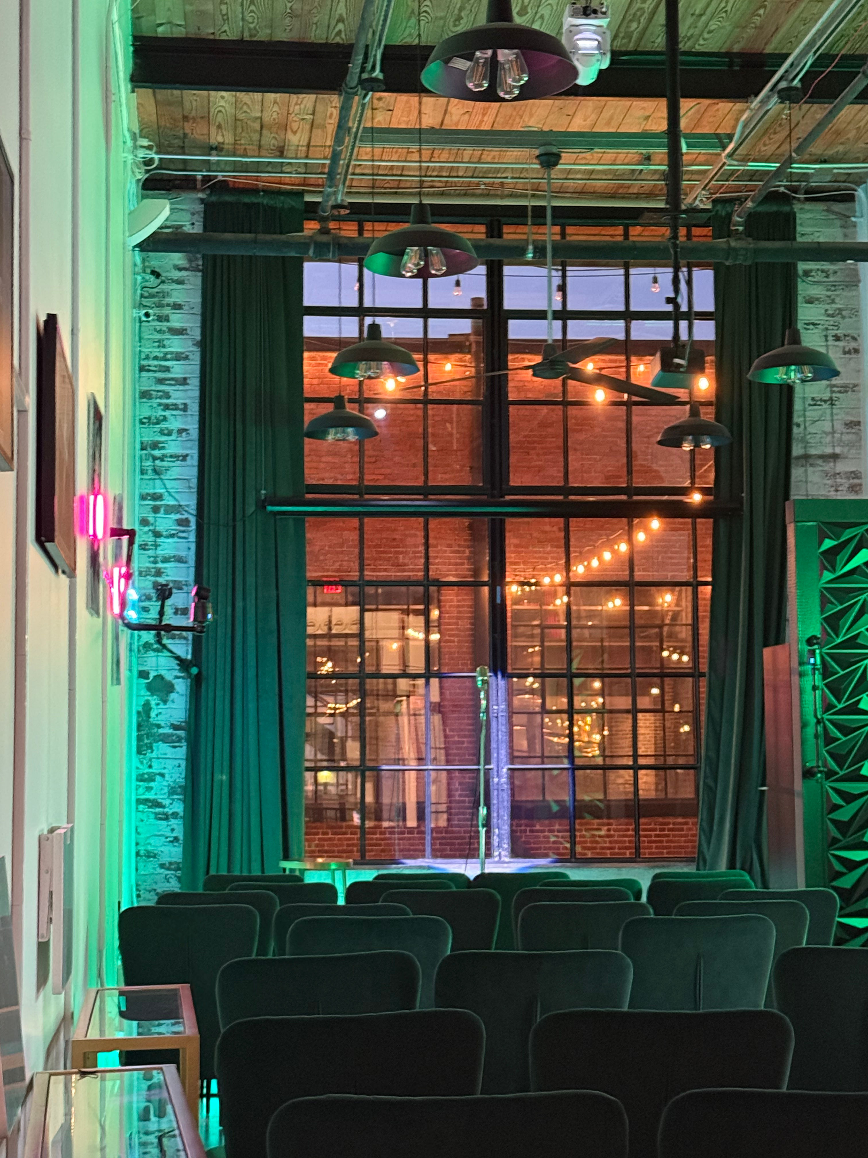 Industrial loft event space with rows of chairs facing a small stage and large multi-pane window, exposed brick and hanging pendant lamps, warm string lights outside and green uplighting inside