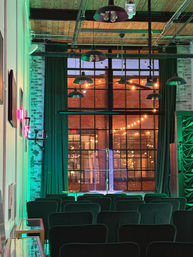 Industrial loft event space with rows of chairs facing a small stage and large multi-pane window, exposed brick and hanging pendant lamps, warm string lights outside and green uplighting inside