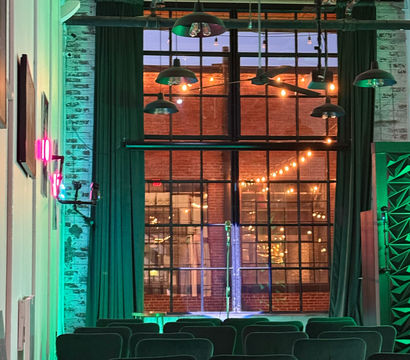 Industrial loft event space with rows of chairs facing a small stage and large multi-pane window, exposed brick and hanging pendant lamps, warm string lights outside and green uplighting inside