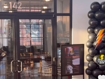 Glass-door entrance to a modern loft-style studio with checkered floor, wooden chairs, industrial windows, and a black balloon column topped with a lightning-bolt decoration and a freestanding poster sign.