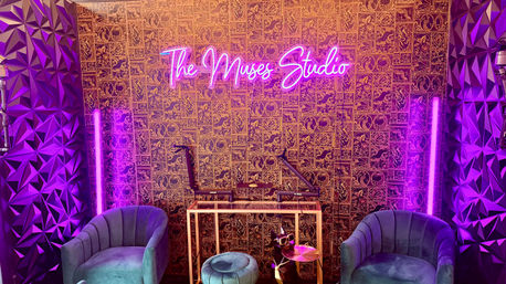 Neon-lit recording studio interior with pink-purple glow, patterned gold wallpaper, teal velvet armchairs, small round ottoman, microphones on adjustable arms, geometric acoustic panels and vertical LED tube lights.