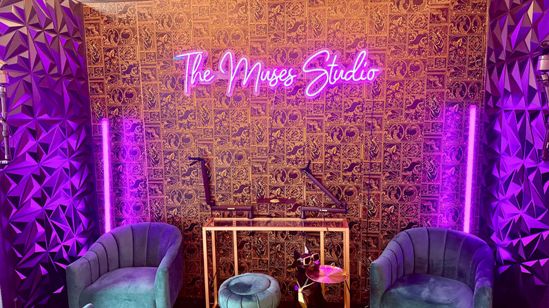 Neon-lit recording studio interior with pink-purple glow, patterned gold wallpaper, teal velvet armchairs, small round ottoman, microphones on adjustable arms, geometric acoustic panels and vertical LED tube lights.