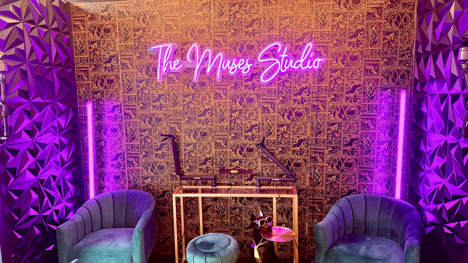 Neon-lit recording studio interior with pink-purple glow, patterned gold wallpaper, teal velvet armchairs, small round ottoman, microphones on adjustable arms, geometric acoustic panels and vertical LED tube lights.