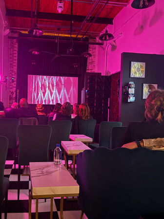 Intimate loft-style screening room bathed in pink light, with rows of upholstered chairs, small marble-top side tables on a black-and-white checkered floor, and a projected abstract film on a front screen.
