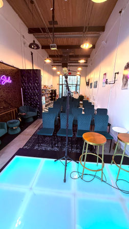 Microphone on a stand centered on a glowing blue stage with two gold stools, facing rows of teal quilted chairs in a cozy industrial studio venue with hanging pendant lights.