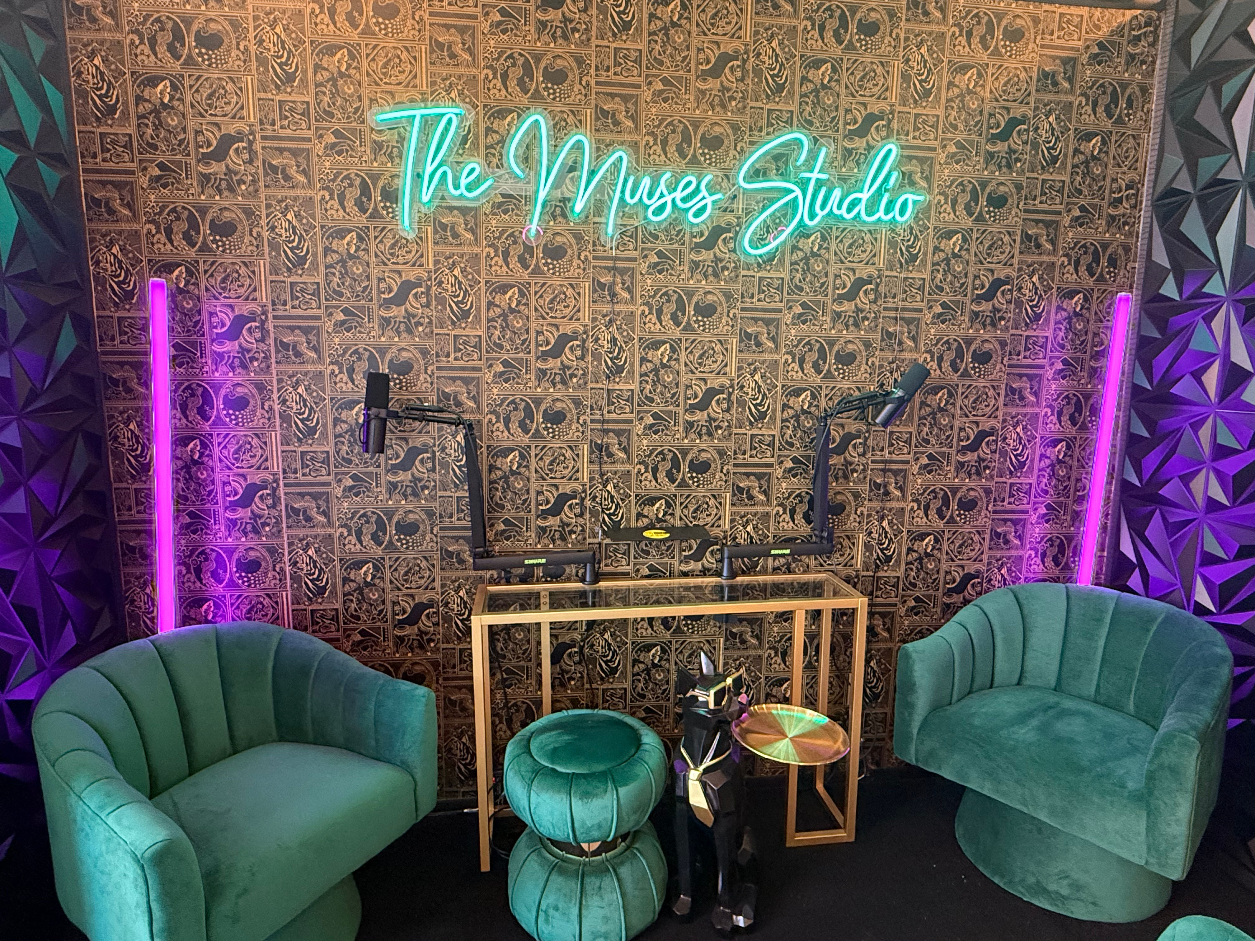 Cozy podcast studio setup with teal neon script sign, patterned decorative wall, two green velvet barrel chairs and ottomans, gold-framed console with dual microphone arms, purple LED light strips and geometric acoustic panels.