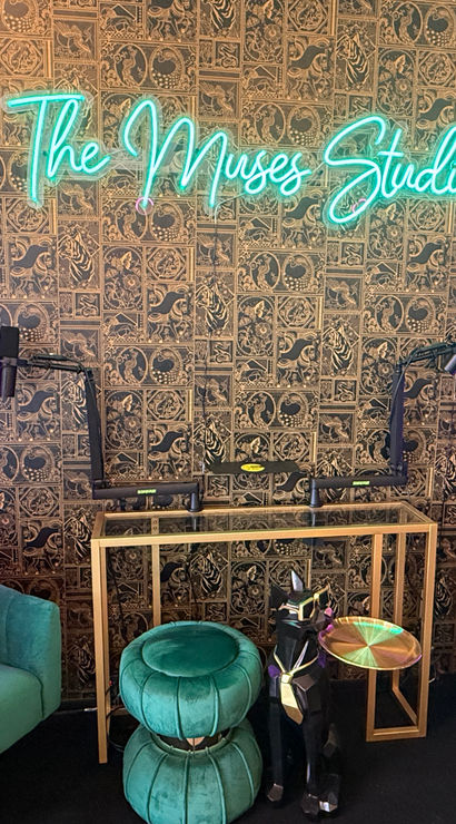 Cozy podcast studio setup with teal neon script sign, patterned decorative wall, two green velvet barrel chairs and ottomans, gold-framed console with dual microphone arms, purple LED light strips and geometric acoustic panels.