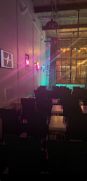 Cozy industrial loft event space with rows of chairs and small tables, pink and teal stage lights on an exposed brick wall, factory-style windows and hanging pendant lights.