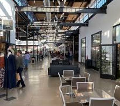 Industrial-style indoor food hall corridor with high exposed-beam ceiling, glass storefronts, metal tables and chairs and lounge seating, people browsing and dining