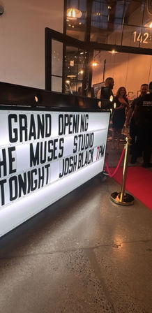 Illuminated marquee announcing a grand opening and performer tonight at a modern studio entrance, with a red carpet, gold stanchion and guests inside.