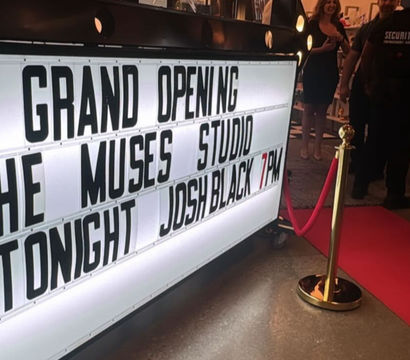 Illuminated marquee announcing a grand opening and performer tonight at a modern studio entrance, with a red carpet, gold stanchion and guests inside.
