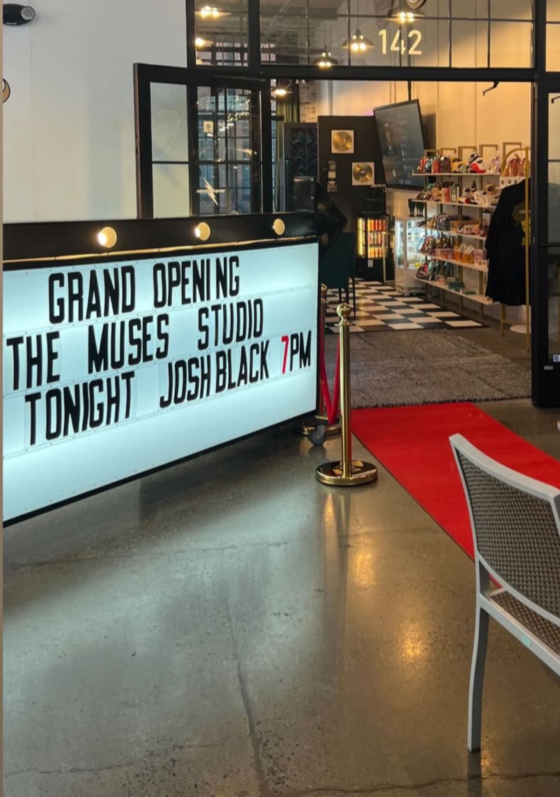 Backlit marquee announcing a grand opening and 7PM event next to a red carpet leading into a modern studio storefront with checkered tile and retail display shelves.