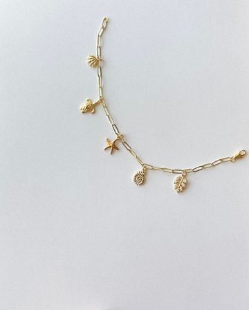 Gold beachy charm bracelet flat lay on light background — delicate chain with shell, turtle, starfish, textured disc and leaf charms, stylish summer jewelry
