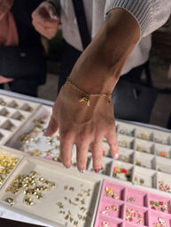 Close-up of a hand with colorful nail art wearing a delicate gold charm bracelet over jewelry display trays filled with small gold and pastel charms