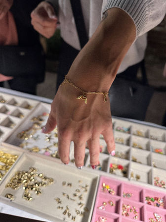 Close-up of a hand with colorful nail art wearing a delicate gold charm bracelet over jewelry display trays filled with small gold and pastel charms