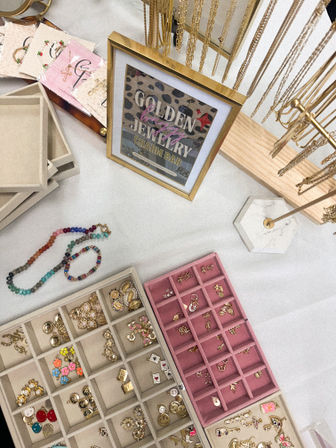 Top-down view of a tabletop jewelry display: gold chain necklaces on a wooden stand next to a framed sign, beige and pink compartment trays of gold charms, colorful beaded bracelets and stud earrings — boutique or craft fair setup.