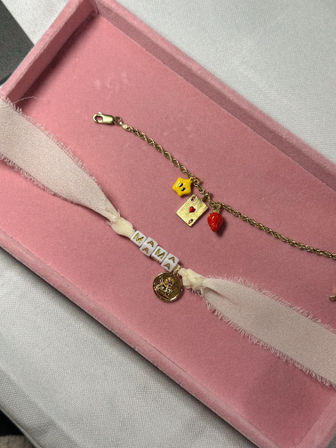 Pink velvet jewelry tray on light gray fabric holding two bracelets: a gold chain with a yellow star, tiny playing-card charm and red strawberry charm, and a pale-pink ribbon bracelet with white 'MAMA' beads and a small gold coin charm.