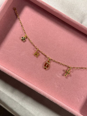 Gold charm necklace with mini football, letter H and cross charms displayed on a pink velvet jewelry tray, close-up flat-lay