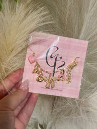 Hand holding a pink gingham jewelry card displaying a gold charm necklace with a pink heart, mini teddy, letter 'S', pearl bow, horseshoe and spiral charm; polka-dot manicure and fluffy pampas grass background.
