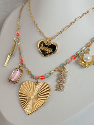 Layered gold necklaces on white display: delicate chain with a black-enamel heart pendant engraved honey, and a colorful beaded chain featuring a large ribbed gold heart pendant, pink crystal, pearl flower, and sparkling rhinestone charm