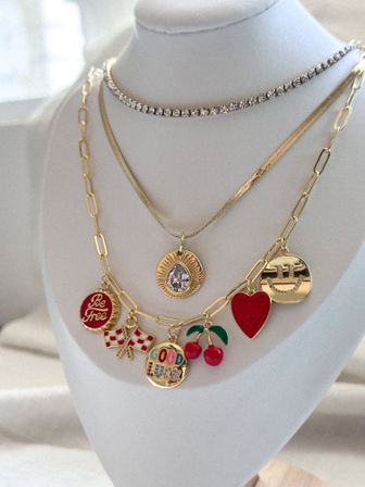Layered gold necklaces on a white display bust: rhinestone choker, gold teardrop pendant and charm chain with red cherries, red heart, smiling face, "Be Free" disc, checkered flags and colorful "Good Luck" medallion.