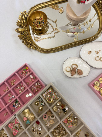 Flatlay jewelry display on a white table: ornate gold mirror tray with necklaces and a tiny bowl of colorful beads, ceramic dishes with rings, and pink and beige compartment trays filled with gold earrings and charms.