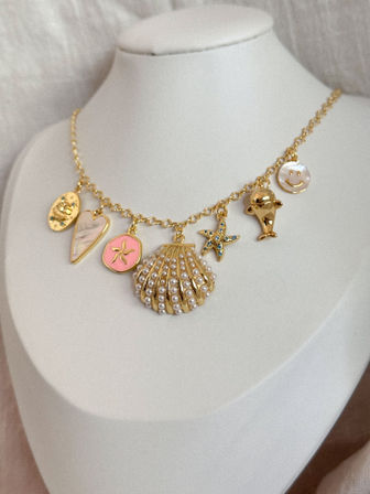 Playful coastal-inspired gold charm necklace with pearl-studded seashell, starfish, heart, smiley and dolphin charms displayed on a white bust — beachy summer jewelry