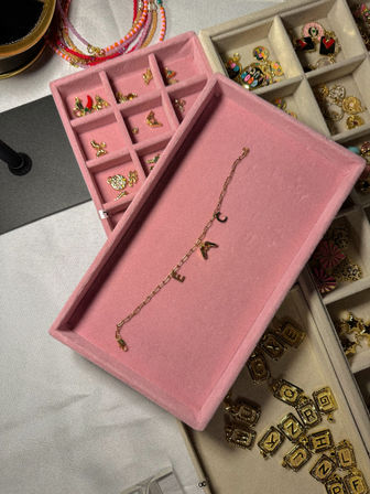 Close-up of a pink velvet jewelry tray on a white vanity holding a delicate gold chain bracelet with letter charms, surrounded by compartmented organizer sections of gold letter charms, enamel charms and colorful beaded necklaces.