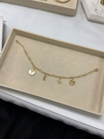 Gold charm bracelet on a beige jewelry tray — delicate chain with a palm coin, tiny bar, airplane charm and textured heart, stylish jewelry display