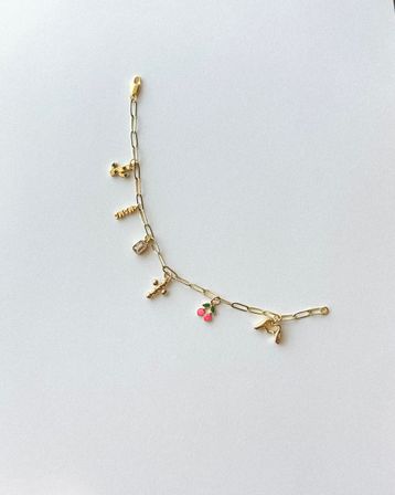 Gold link charm bracelet flat-lay on white background featuring a cactus charm, 'MAMA' bar, small crystal, bead cluster, pink enamel cherries and a gold high-heel charm