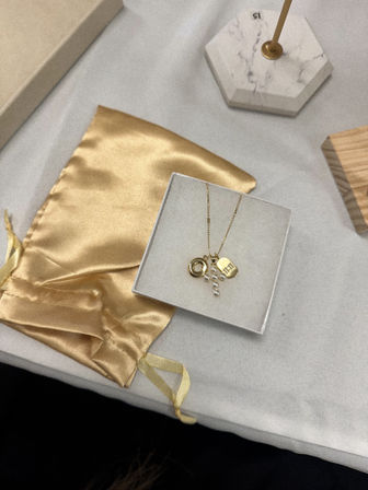 Jewelry flatlay: gold necklace with pearl cluster and engraved tag in a white gift box on a light gray surface, beside a gold satin pouch and marble lamp base
