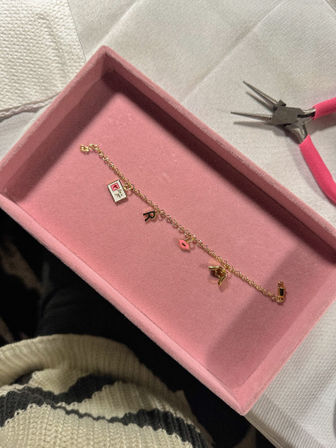 Gold charm bracelet with tiny book, letter R, pink lips and bird charms displayed in a pink velvet jewelry tray next to pink-handled pliers on a white cloth workspace