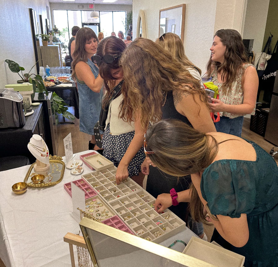 Women browsing trays of handmade jewelry at a bright indoor pop-up boutique, rings and necklaces on display, casual summer shoppers.