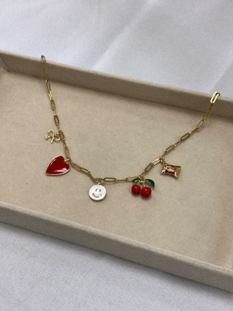 Playful gold paperclip-chain charm necklace with red enamel heart, smiling face, cherries and red-accent rectangular charm resting in a beige jewelry tray