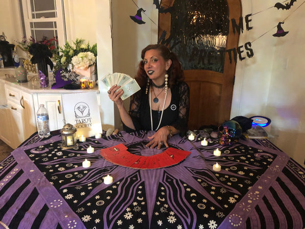 Tarot reader at a Halloween-themed indoor event seated behind a purple star-and-moon tarot cloth, holding fanned cards with a red spread on the table, surrounded by flickering tea lights, decorative skull, flowers and witch-hat garlands.