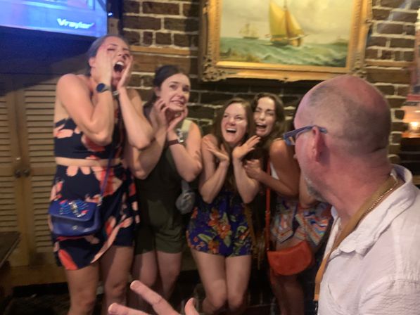 Four women playfully posing with exaggerated shocked expressions and laughter against a brick wall with a framed sailboat painting, while a man in glasses watches in a lively indoor bar setting — night out vibe.