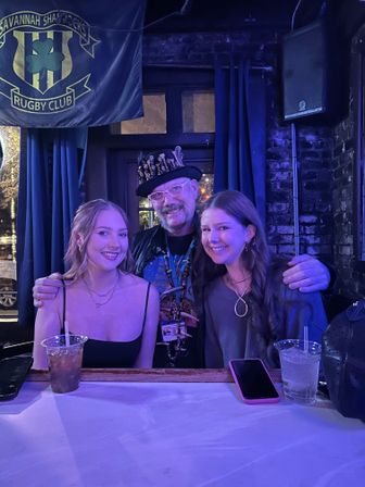 Three smiling people at a blue-lit bar in Savannah — two young women seated with drinks and a man wearing a decorative studded top hat, posed in front of exposed brick and a rugby-style banner.