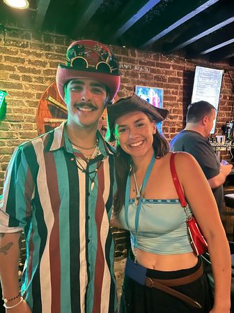 Smiling patrons wearing a steampunk top hat with goggles and a pirate-style hat pose in a brick-walled downtown pub by beer taps during a lively night out.