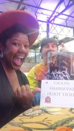 Selfie of two excited people at an indoor historic event space — woman in a wide-brim hat and man in a bandana holding a ghost-tour brochure beneath exposed metal beams.