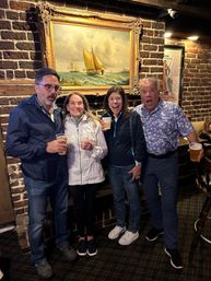 Four adults laughing and holding plastic cups of beer in a cozy brick-walled pub beneath a large framed sailboat painting, casual night out vibe.