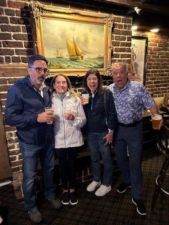 Four adults laughing and holding plastic cups of beer in a cozy brick-walled pub beneath a large framed sailboat painting, casual night out vibe.