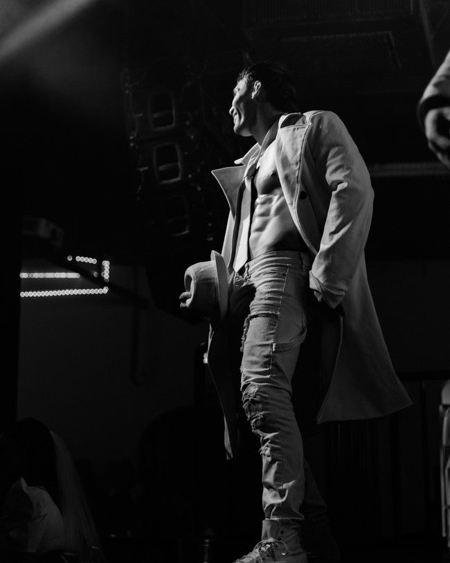 Black-and-white photo of a shirtless stage performer on a nightclub stage under a spotlight, wearing a trench coat and ripped jeans, holding a hat and smiling
